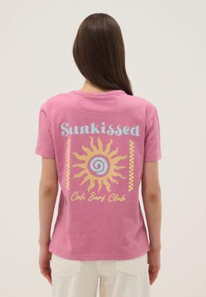 Donna con lunghi capelli castani indossa una maglietta rosa con grafica di sole giallo e testo blu "Sunkissed Cali Surf Club", abbinata a pantaloni bianchi.