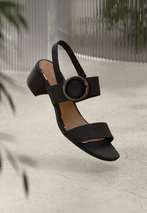 Schwarze Wildleder-Absatzsandalen mit zwei breiten Riemen, kreisförmigem metallic Akzent, niedrigem Blockabsatz und offener Zehenpartie vor einem strukturierten Hintergrund.