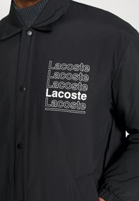 Černá bunda s límečkem, na které se opakuje bílé text "Lacoste." Materiál vypadá hladce s lesklým povrchem.