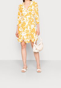 Robe florale jaune à manches longues, décolleté en V et ourlet à volants. Associée à un sac à main blanc avec une anse en perles et des sandales à bout ouvert.