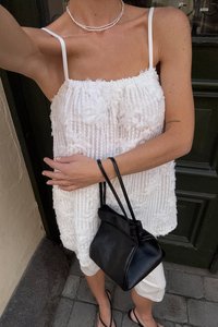 Witte textuurtop met bloemdetails, gecombineerd met een zwarte leren handtas en lichte shorts; heeft dunne bandjes en een gelaagd ontwerp.