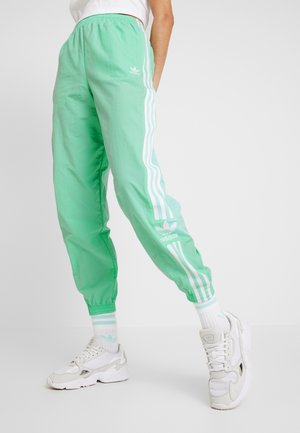 Person iført mintgrønne Adidas træningsbukser med hvide striber, kombineret med hvide sneakers og sokker, står foran en ensfarvet baggrund.
