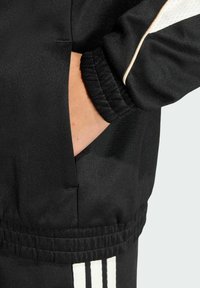 Čierna športová bunda s elastickými manžetami a pásom, s bočnou kapsou a kontrastnými bielymi pruhmi pozdĺž rukávov.