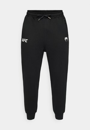 Črne trenerke iz mehke tkanine, z elastičnim pasom z vrvico in belimi logotipi na straneh, z zategnjenimi gležnji.