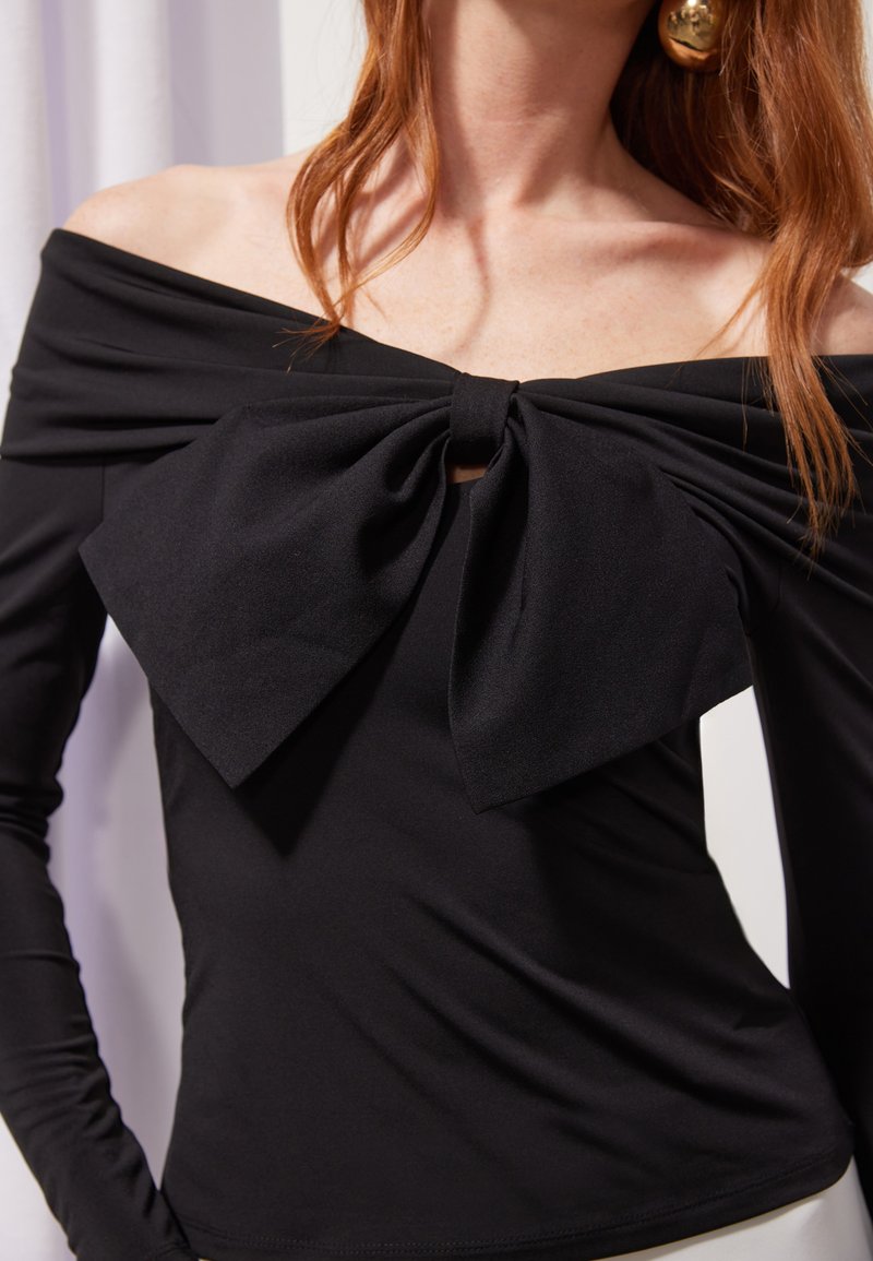 Schwarzes Langarmoberteil mit einem Off-Shoulder-Design, das vorne mit einer großen Schleifenakzentuierung versehen ist und einen glatten, dehnbaren Stoff hat.