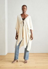 Lysinbeige kimono-inspirert cardigan med knytelukking, løs passform, korte ermer og en luftig design, båret over en mørk topp og jeans.
