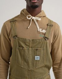 Salopette rayée verte et jaune avec une poche avant, dotée de quincaillerie en argent et d'une étiquette Lee Crayola, portée sur un sweat à capuche marron.