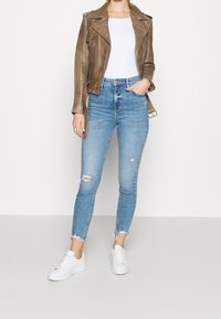 Giubbotto in pelle marrone da biker, camicia bianca attillata, jeans skinny strappati blu chiaro con orlo sfilacciato e sneakers bianche.