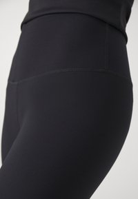 Höga svarta leggings gjorda av slätt, stretchigt tyg med platta sömmar och en åtsittande silhuett. Texturen är slät och mjuk att ta på.