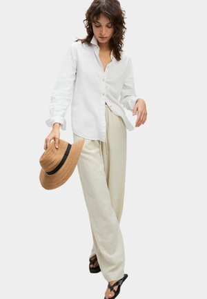 Chemise blanche à manches retroussées, pantalon large crème, et chapeau en paille marron avec un bandeau noir tenu à la main. Tongs noires.