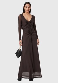 Langes, braunes Kleid mit tiefem V-Ausschnitt, gerafften Details vorne, transparenten langen Ärmeln und bodenlangem Schnitt. Schwarze Tasche mit Kette.