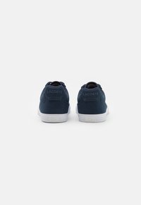 Marineblå kanvas-sneakers med hvite gummisåler og teksturerte detaljer, med en subtil preget logo på hælen. Klassisk lave design.
