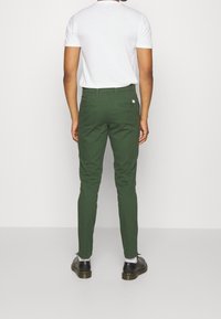 Zielone bawełniane chinosy z dwoma tylnymi kieszeniami, dopasowanym krojem i kontrastowymi metalowymi elementami. W zestawie z białym t-shirtem z krótkim rękawem i czarnymi butami.