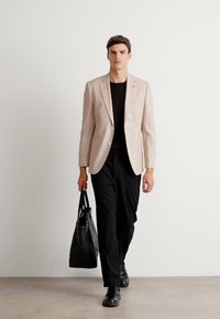 Svetloružový blazer, čierna košeľa, čierne nohavice a čierne topánky. Model nesie čiernu tašku na rameno. Hladká tkanina, štruktúrovaný dizajn.