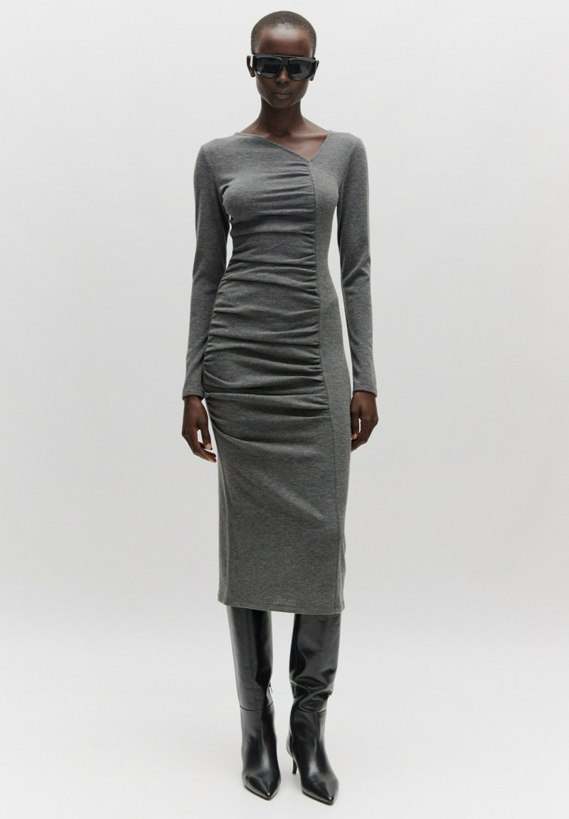 Langärmliges, graues Kleid mit gerafftem Seiten-Detail, figurbetontem Schnitt und Midilänge. Kombiniert mit schwarzen, hochhackigen, spitzen Stiefeln.