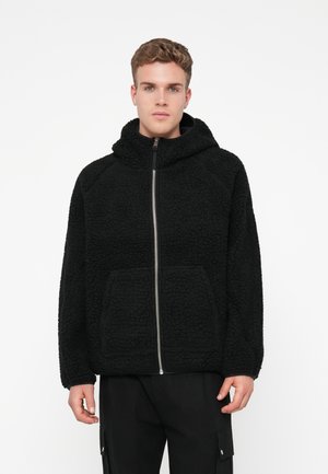 Musta vetoketjuhuppari, joka on valmistettu teksturoidusta fleece-materiaalista. Siinä on suuri huppu ja etutaskut. Mallissa on rento istuvuus ja raglan-hihat.