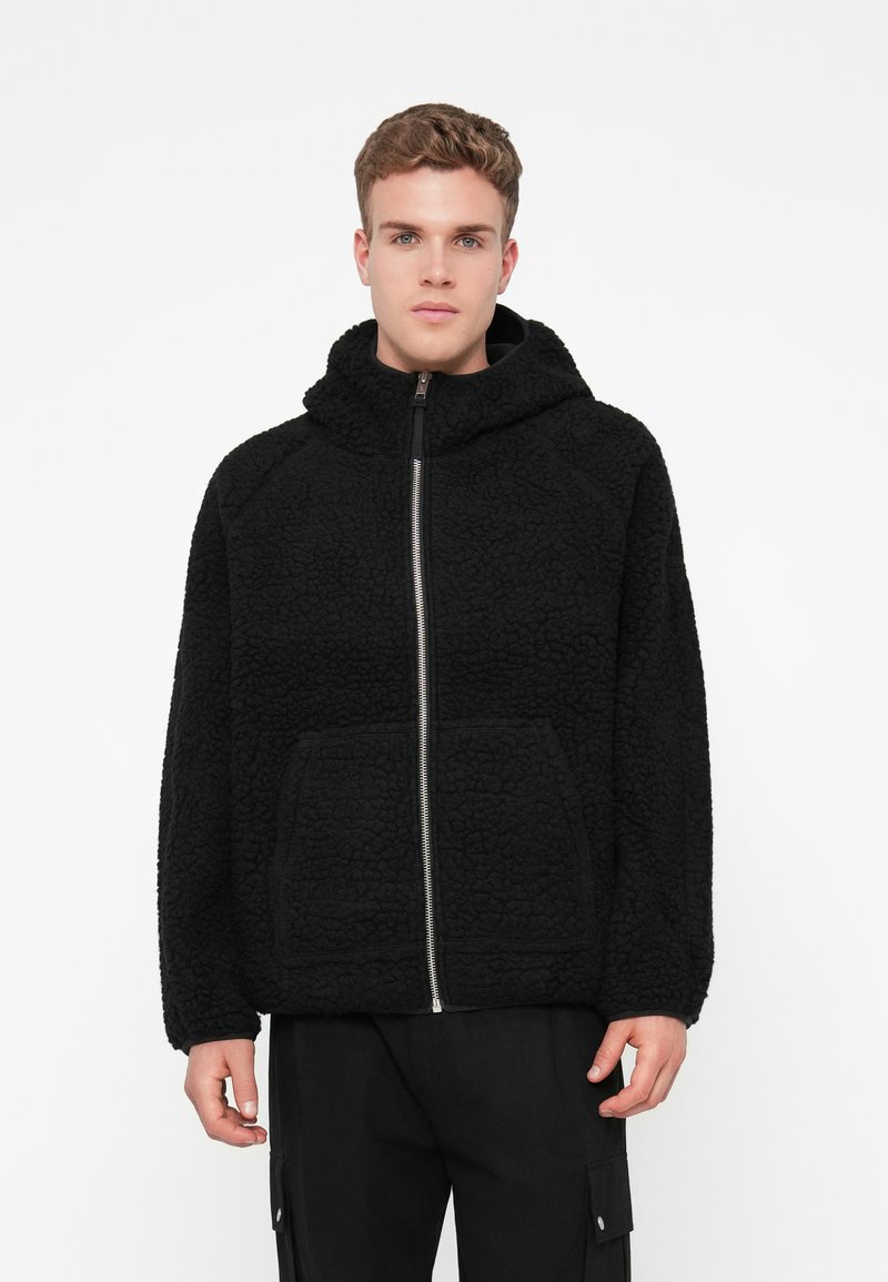 Sweatshirt preto com fecho, feito de fleece texturizado, apresentando um grande capuz e bolsos frontais. O design tem um corte descontraído com mangas raglan.