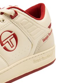 Béžové tenisky s červenými akcentmi, logom a textúrovaným povrchom; obsahujú šesť ventilačných otvorov blízko šnúrok a kontrastnú gumovú podrážku.