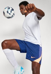 Biela tričko s odvodom vlhkosti, modré šortky s oranžovým logom Nike, biele ponožky po lýtka a svetlomodré kopačky na futbal. Čierny futbalový loptu v pohybe.