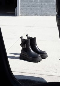 Paar schwarze klobige Plateau-Stiefeletten mit Schnallendetail auf hellem Betonboden in der Nähe einer grauen Ziegelwand.