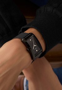 Schwarze rechteckige Uhr mit einem strukturierten Netzarmband. Verfügt über ein schwarz-farbenes Ziffernblatt mit silbernen Zeigern und Markierungen, auf dem der Markenname "Guess" angezeigt wird.