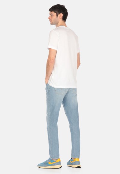 T-shirt bianca a maniche corte, jeans azzurri chiari, sneakers blu e gialle. La vista posteriore mette in evidenza la vestibilità casual e il morbido materiale di cotone.
