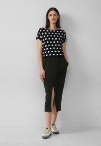 Zwarte top met korte mouwen en witte stippen, gecombineerd met een zwarte rok tot de knie met een split aan de voorkant. Witte en beige sneakers.