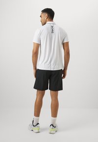 Camisa polo branca com uma impressão vertical preta "BORG" nas costas, combinada com calções pretos, usada por uma pessoa de costas.