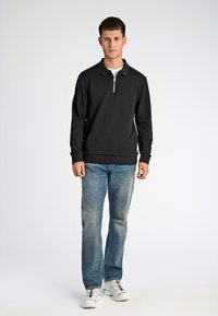 Felpa nera con colletto zip, indossata sopra una camicia bianca; abbinata a jeans azzurri e sneakers bianche. Sfondo semplice.
