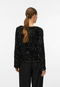 Svart paljett-topp med långa ärmar, strukturerad yta, avslappnad passform, rund halsringning, ihop med svarta vida byxor.