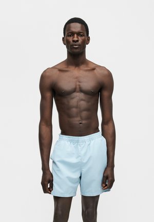 Mężczyzna z ciemną skórą bez koszulki, ubrany w jasnoniebieskie kąpielówki, stoi zwrócony przodem na gładkim jasnym tle.