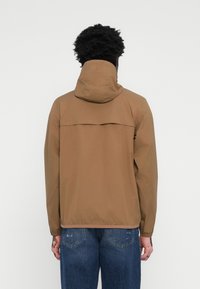 Brown lightweight jacket with a hood, featuring a cross-back design, elastic cuffs, and a plain texture. Paired with blue denim jeans.