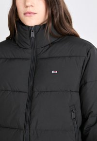 Svart puffjakke med høy krage, horisontale sømmer, frontlås og en liten logo på brystet.