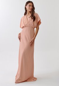 Long, flowing, peach-colored dress made from smooth fabric. Features a deep V-neck, short dolman sleeves, and gathered waist for a draped effect.