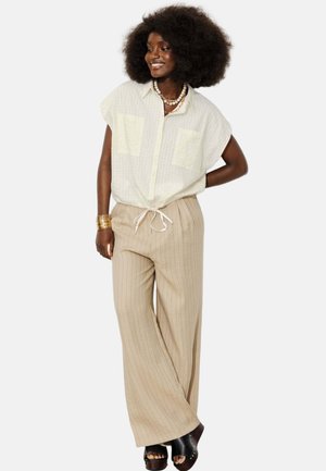 Femme souriante aux grands cheveux bouclés portant un chemisier blanc ample à manches courtes, un pantalon beige à larges rayures, des sandales à plateformes noires et des bijoux en or.