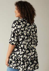 Tunic nera floreale con margherite bianche dai centri gialli, maniche corte, orlo arricciato e un design ampio e fluido.