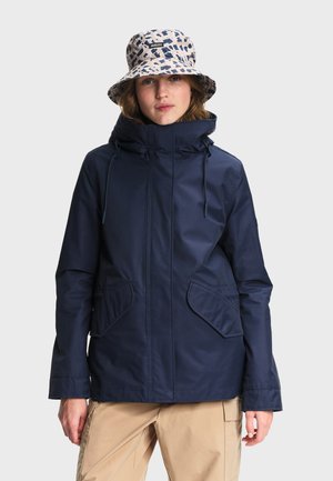 Jeune femme portant une veste à capuche bleu marine, un pantalon beige et un chapeau style seau blanc avec un motif abstrait bleu marine.