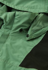 Green jacket with a smooth textile finish, featuring black accents, snap closures, and folded collar design. Visible stitching details.