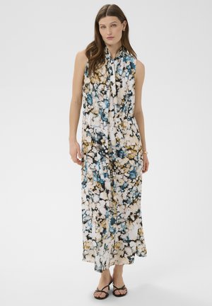 Kobieta w bezrękawowej sukience do kostek z wiązaniem pod szyją, z abstrakcyjnym kwiatowym wzorem w kolorach niebieskim, białym, czarnym i złotym oraz w czarnych sandałach na obcasie z paskami.