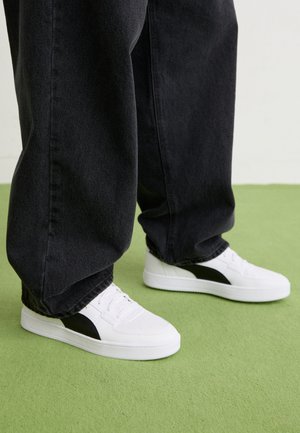 Fehér sportcipők fekete részletekkel, sima szintetikus anyagból, fűzős kialakítással és kerek orral, sötét, bő szabású farmerral párosítva.