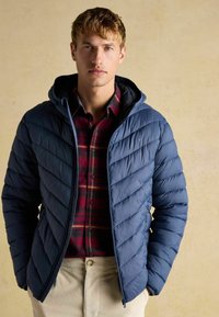 Blå puffjakke med hætte, chevron-dækning og lynlås, lagd over en rød og marineblå ternet skjorte, båret med beigefarvede bukser.