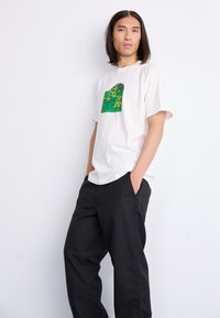 White cotton t-shirt featuring a green graphic print, paired with loose-fitting black trousers. The t-shirt has a classic crew neck design.
