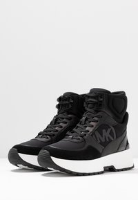 Černé kotníkové tenisky z broušené kůže a syntetických materiálů, s tlustou bílou podrážkou a výrazným logem MK na boku.