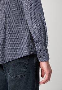 Personne portant une chemise bleu marine à motifs à manches longues et un jean foncé, montrant l’arrière du corps de la taille à la main.
