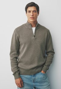 Jonge man met kort donker haar, gekleed in een bruine zip-up sweater en lichtblauwe spijkerbroek, staande tegen een effen lichtgrijze achtergrond.