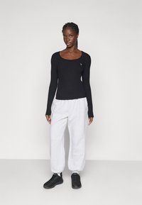 Woman standing against plain background wearing black long-sleeve top, light gray sweatpants, and black sneakers.