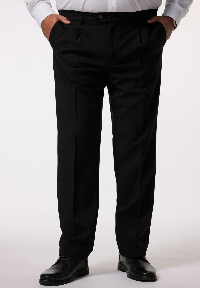 Mężczyzna w czarnych formalnych spodniach z zakładkami, czarnych eleganckich butach, białej koszuli włożonej do środka, z rękami w kieszeniach, stojący na jasnym tle.