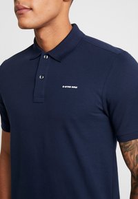 Man wearing navy blue G-Star Raw polo shirt with a two-button placket, showing a tattooed forearm against a plain background.