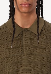 Polo shirt en maille verte avec un design texturé, doté d'une patte de boutonnage et d'un col côtelé contrastant. Vue en gros plan, recadrée au niveau du cou.