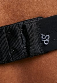 Zwarte verstelbare band gemaakt van ribbelstof, met metalen haakjes en een klein geborduurd logo in grijs. Weergegeven tegen de huid.